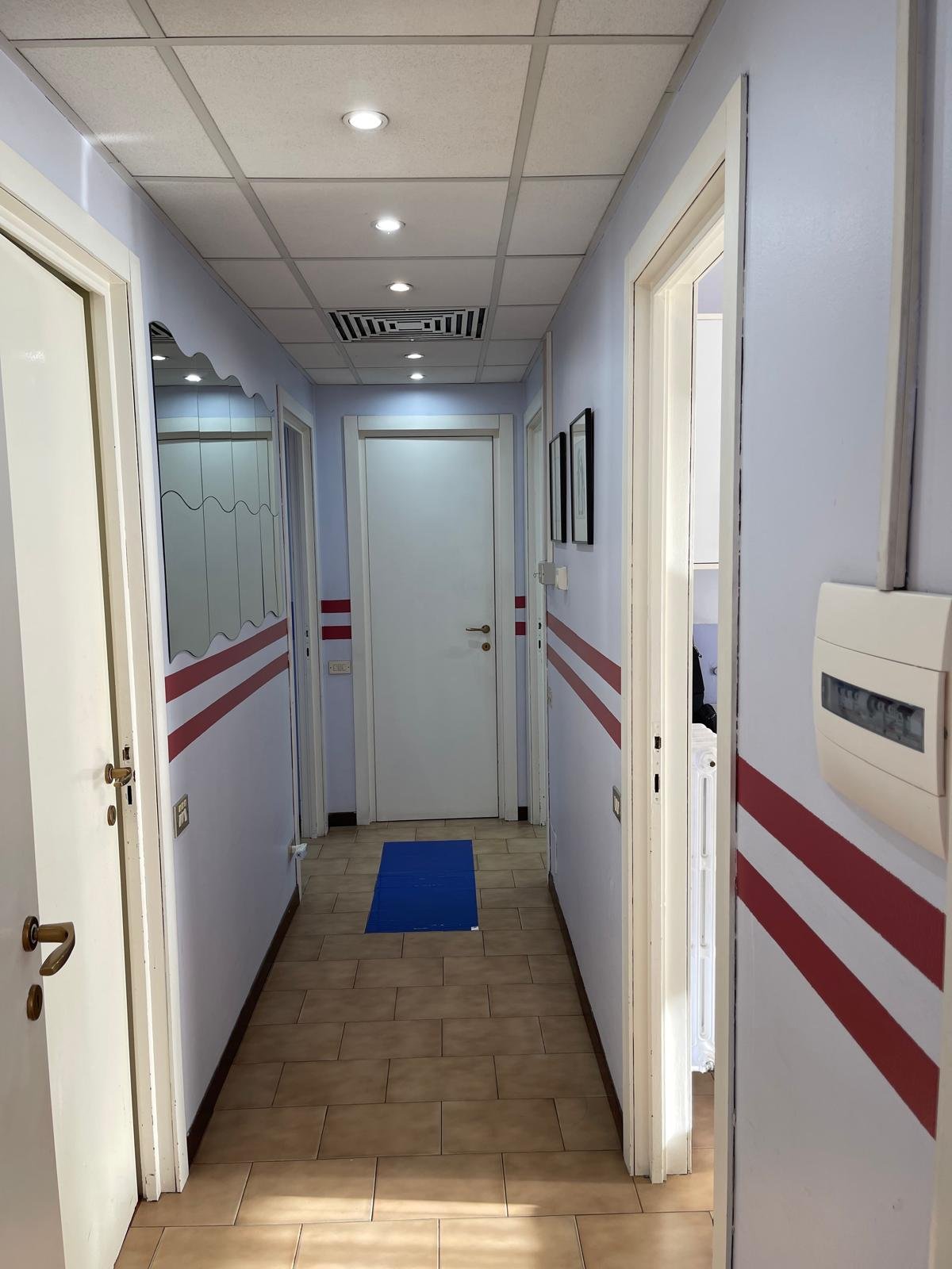 Corridoio interno di un edificio con pareti azzurre, una mobilia a specchio sulla sinistra e tre quadri neri appesi sulla destra.