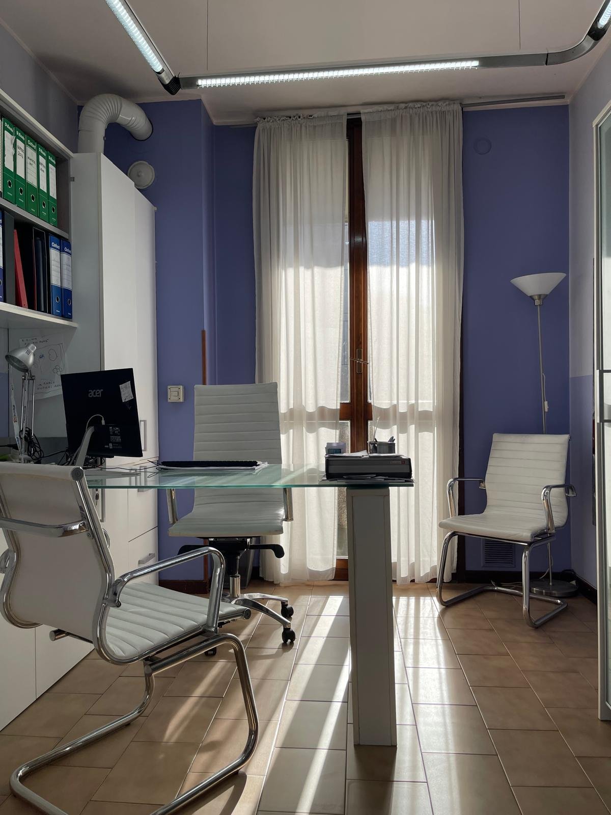 Ufficio con scrivania in vetro, sedia girevole, due sedie bianche con braccioli, mensole con cartelle, computer e lampade, pareti azzurre e finestra con tende bianche.